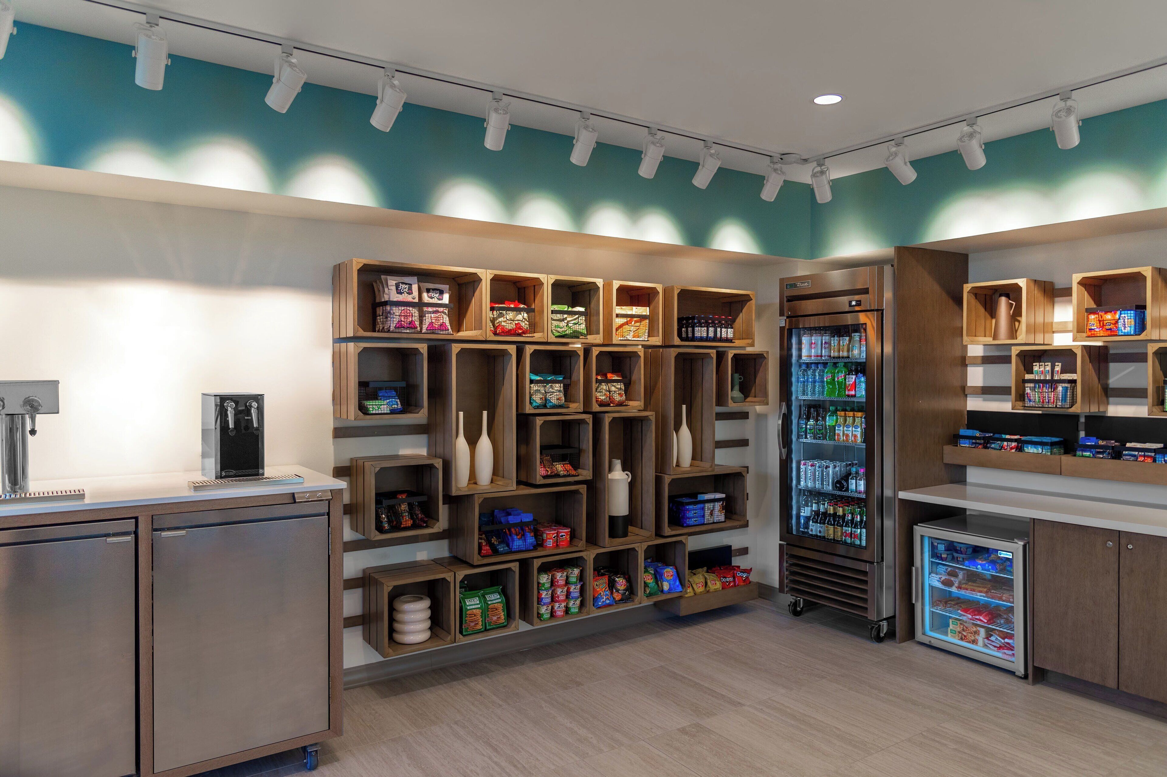 A modern kitchenette with built-in appliances, wooden shelves stocked with snacks, and a refrigerator.