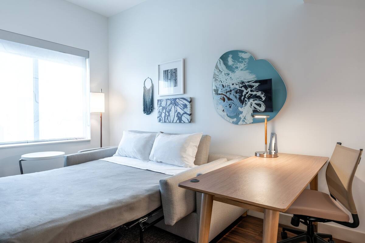 Quarto de hotel moderno com uma cama, uma escrivaninha e uma cadeira. Há obras de arte nas paredes e um abajur.