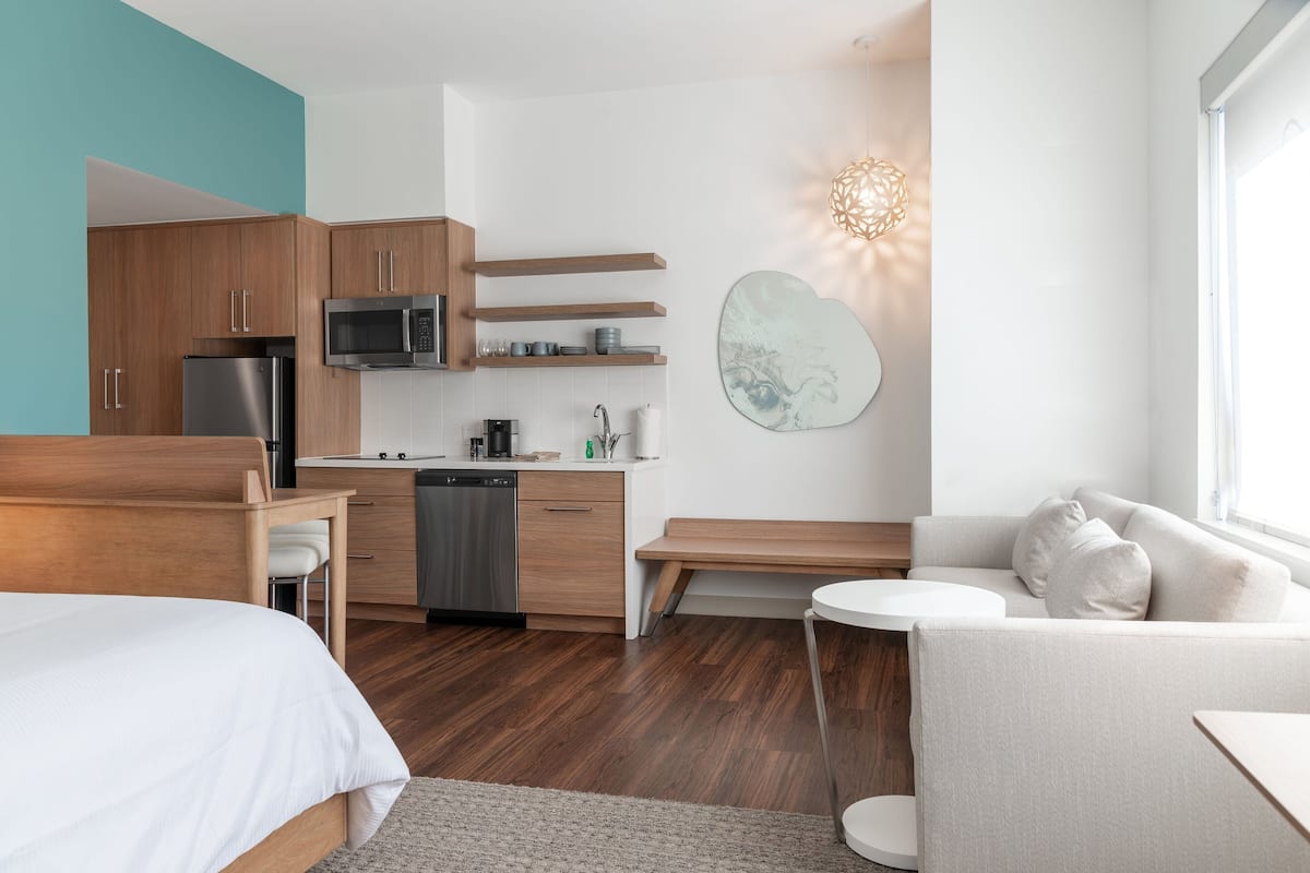Quarto de hotel moderno com cozinha compacta, cama, sofá e uma janela grande.