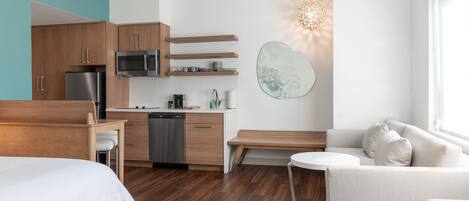 A modern hotel room with a kitchenette, a bed, a sofa, and a large window.