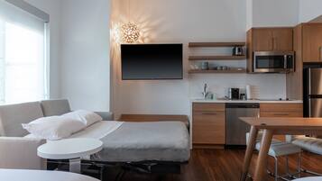 A compact hotel room with a bed, a small dining table, a kitchenette, and a TV mounted on the wall.