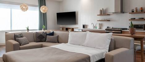 A modern hotel room with a large bed, a sofa, a flat-screen TV, a kitchen area, and a dining table.