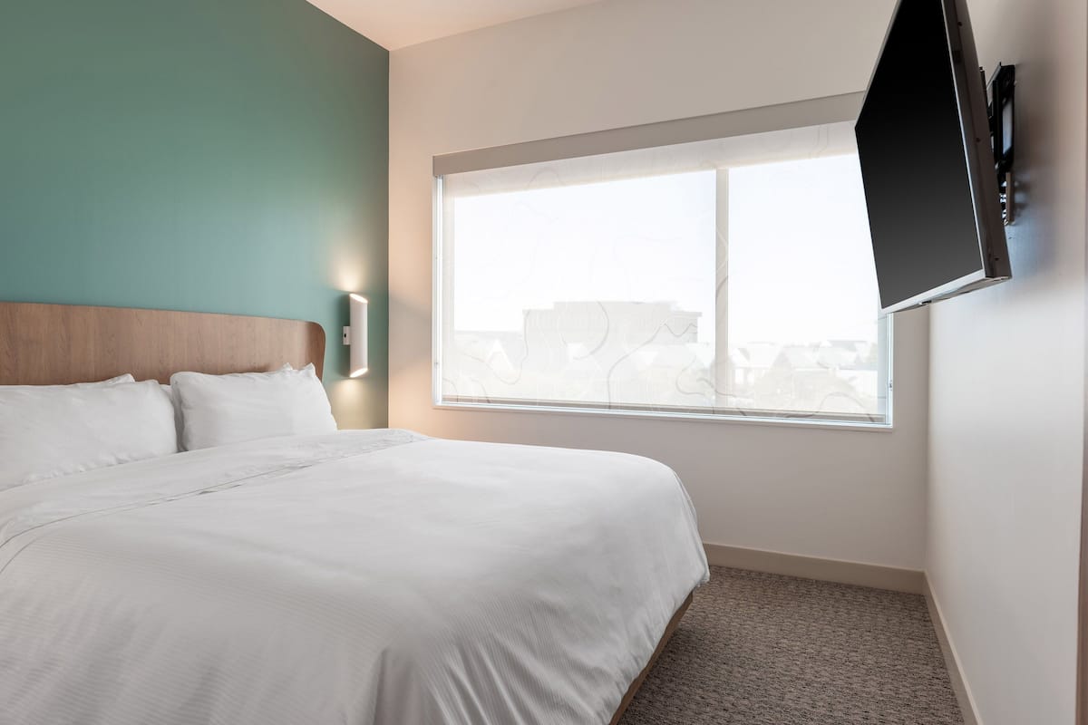Quarto de hotel com uma cama grande, uma janela com persianas, uma televisão de tela plana na parede e uma pequena mesa de cabeceira.