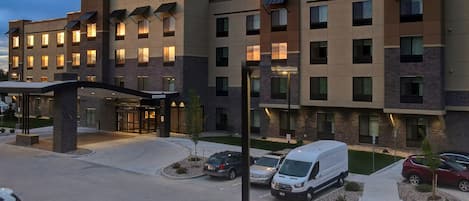 A multi-story hotel with a modern design, featuring a prominent entrance with a covered walkway, several parking spaces, and a well-lit facade.