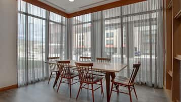 A dining area with a large table and six chairs, large windows with sheer curtains, and a tiled floor.