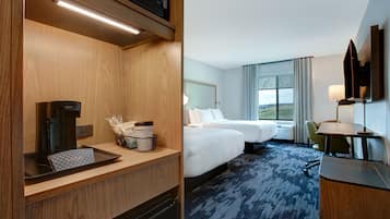 A hotel room with a wooden desk, a coffee maker, a basket with toiletries, a bed, a desk with a lamp, and a view of the outdoors.