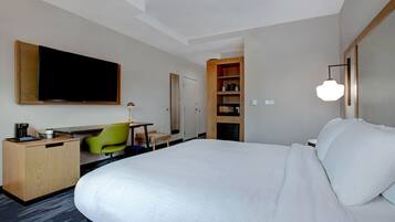 A hotel room with a large bed, a desk with a chair, a television, a bookshelf, and a lamp.