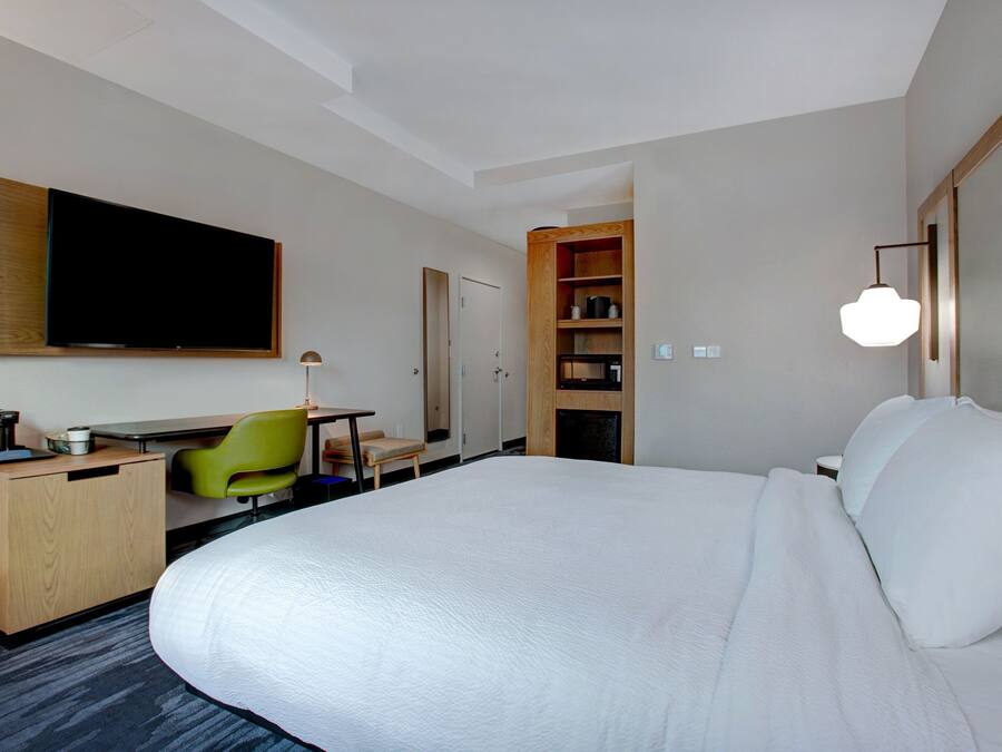 A hotel room with a large bed, a desk with a chair, a television, a bookshelf, and a lamp.
