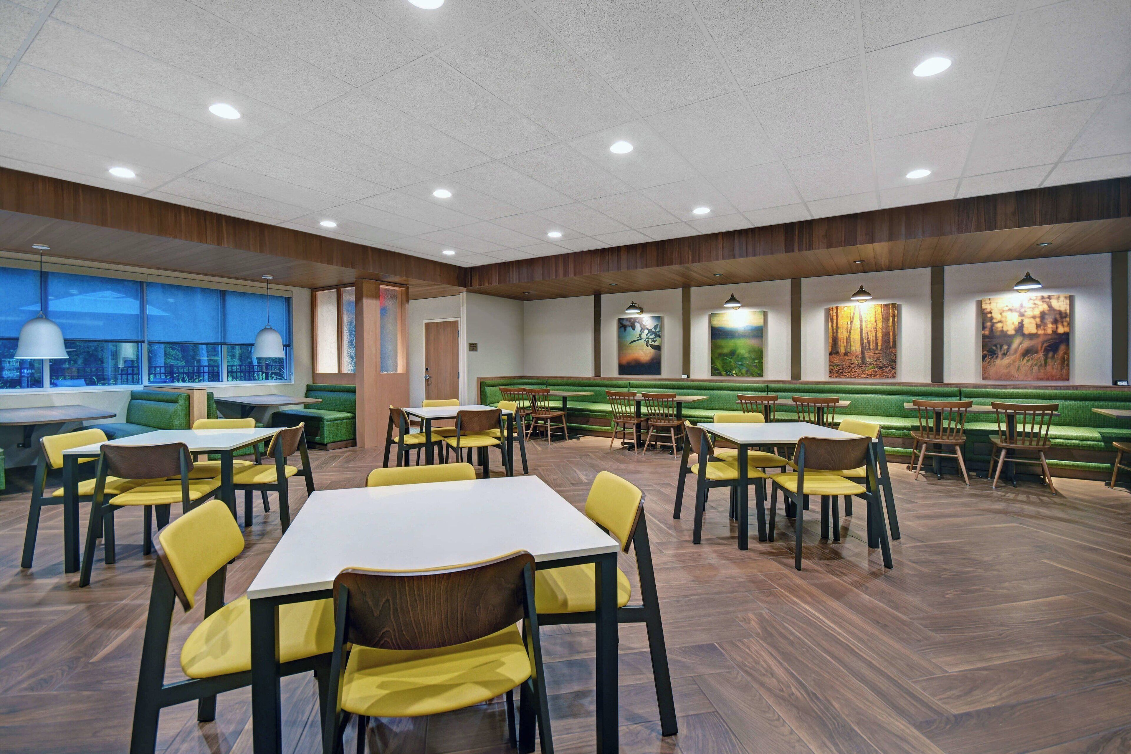A spacious dining area with multiple tables and chairs, green cushioned seating, and artwork on the walls.
