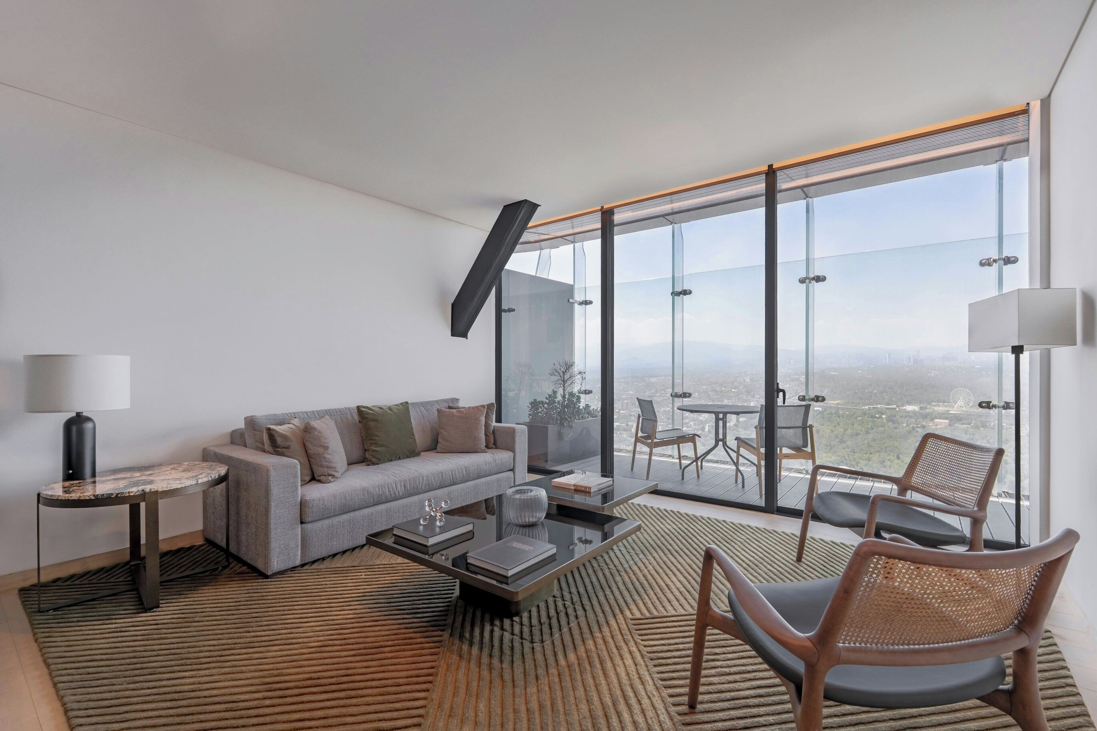 Una sala de estar moderna con sofá, mesa de centro y sillas, ventanas amplias con vista a la ciudad y un balcón con mesa y sillas.