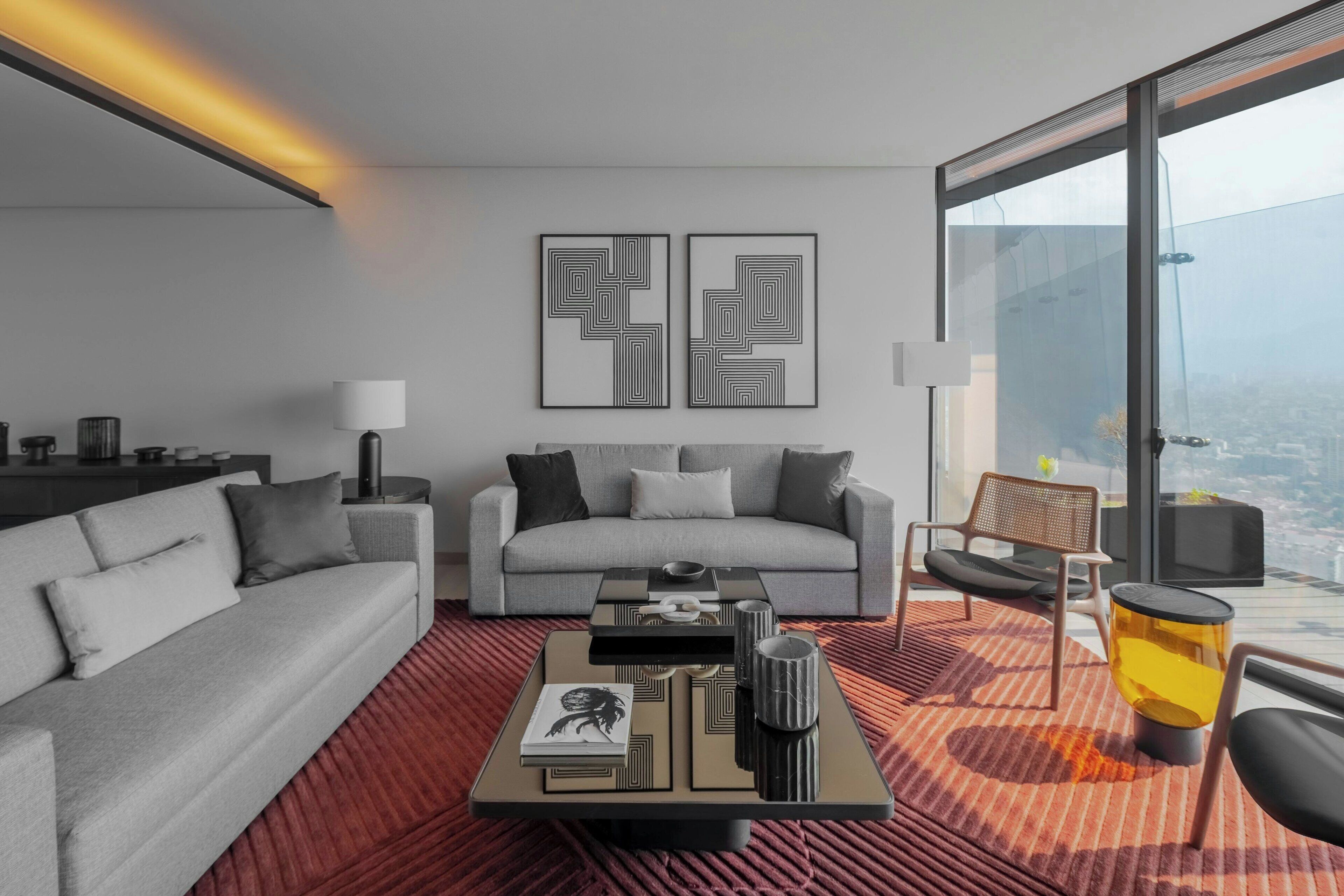 Una sala de estar moderna con un sofá gris, una mesa de centro con libros y objetos decorativos, una alfombra estampada roja y dos piezas de arte abstracto en la pared.