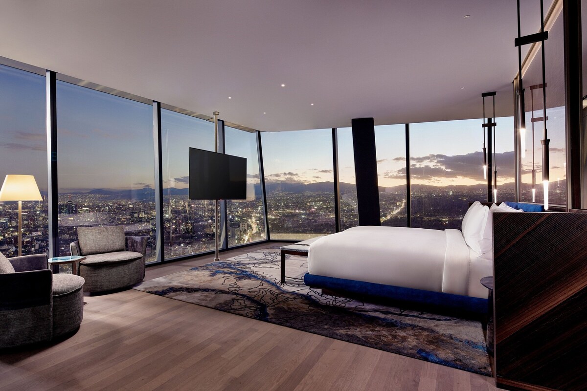 A modern hotel room with a large bed, a TV, and a city view.