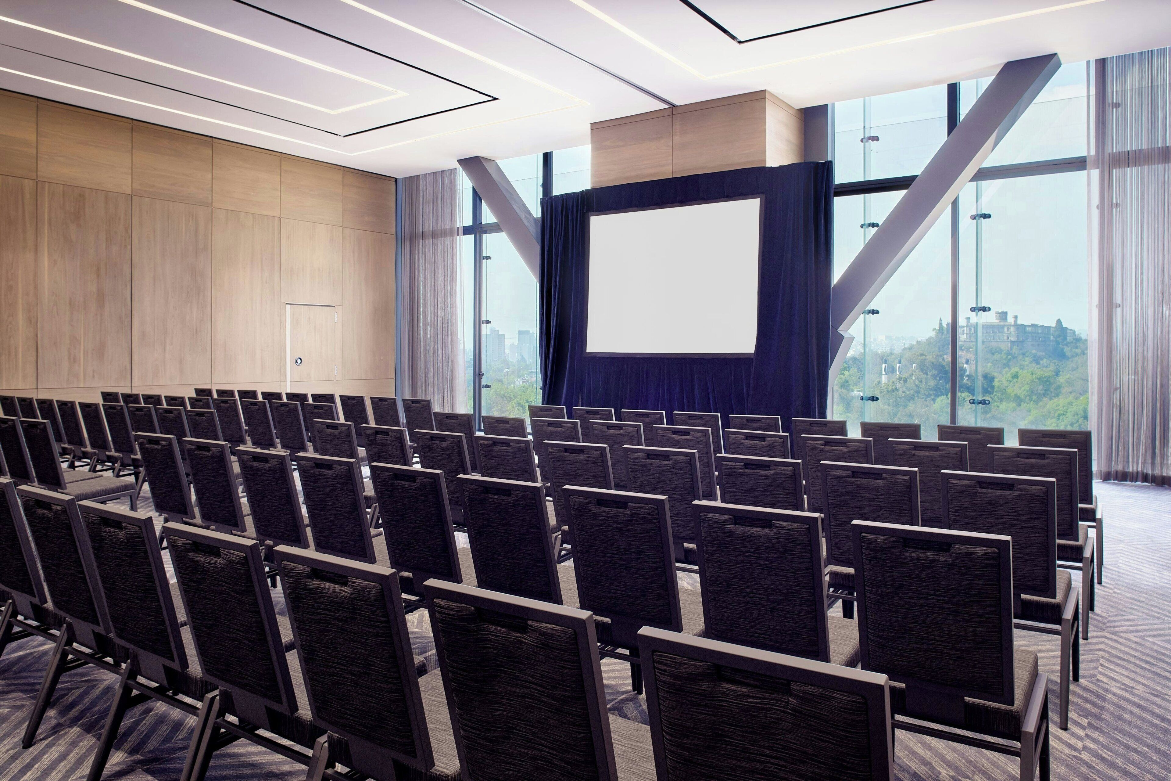 Une salle de conférence avec des rangées de chaises, un grand écran et une vue sur la ville grâce à de grandes fenêtres.