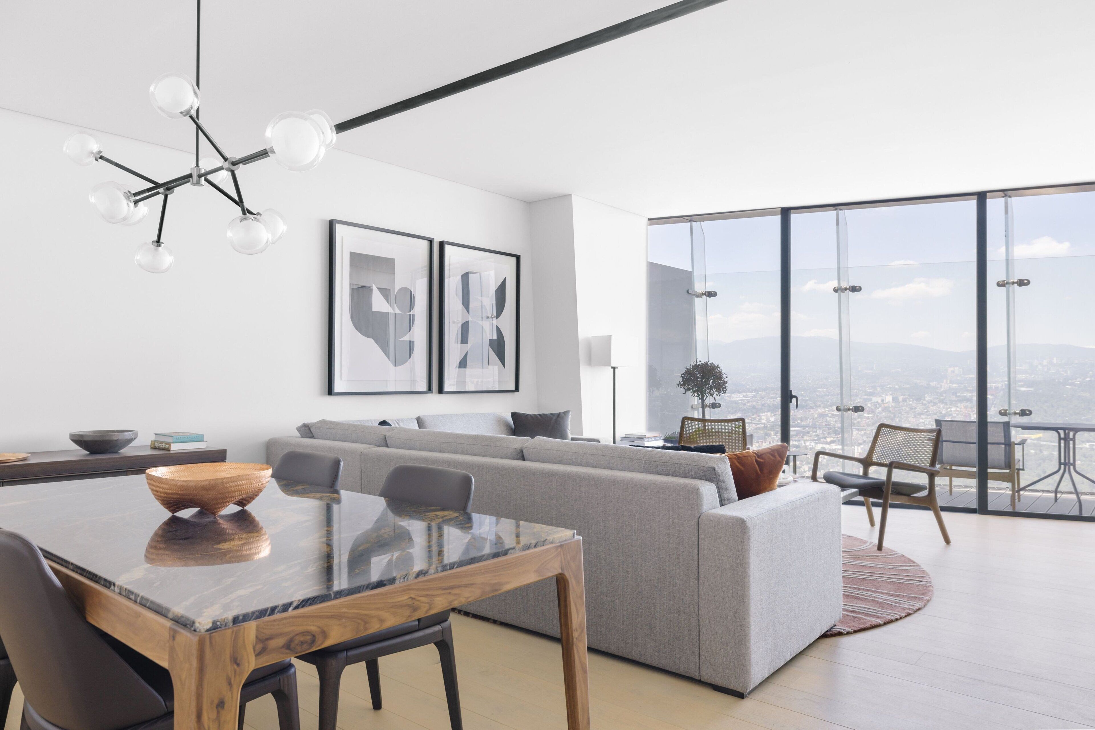 Un salon moderne avec une table à manger en verre, un canapé gris et une vue sur la ville grâce à de grandes fenêtres.