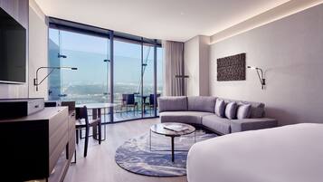 Una habitación de hotel moderna con una cama grande, un sofá, un escritorio con televisor y vistas a un paisaje urbano a través de ventanas de piso a techo.