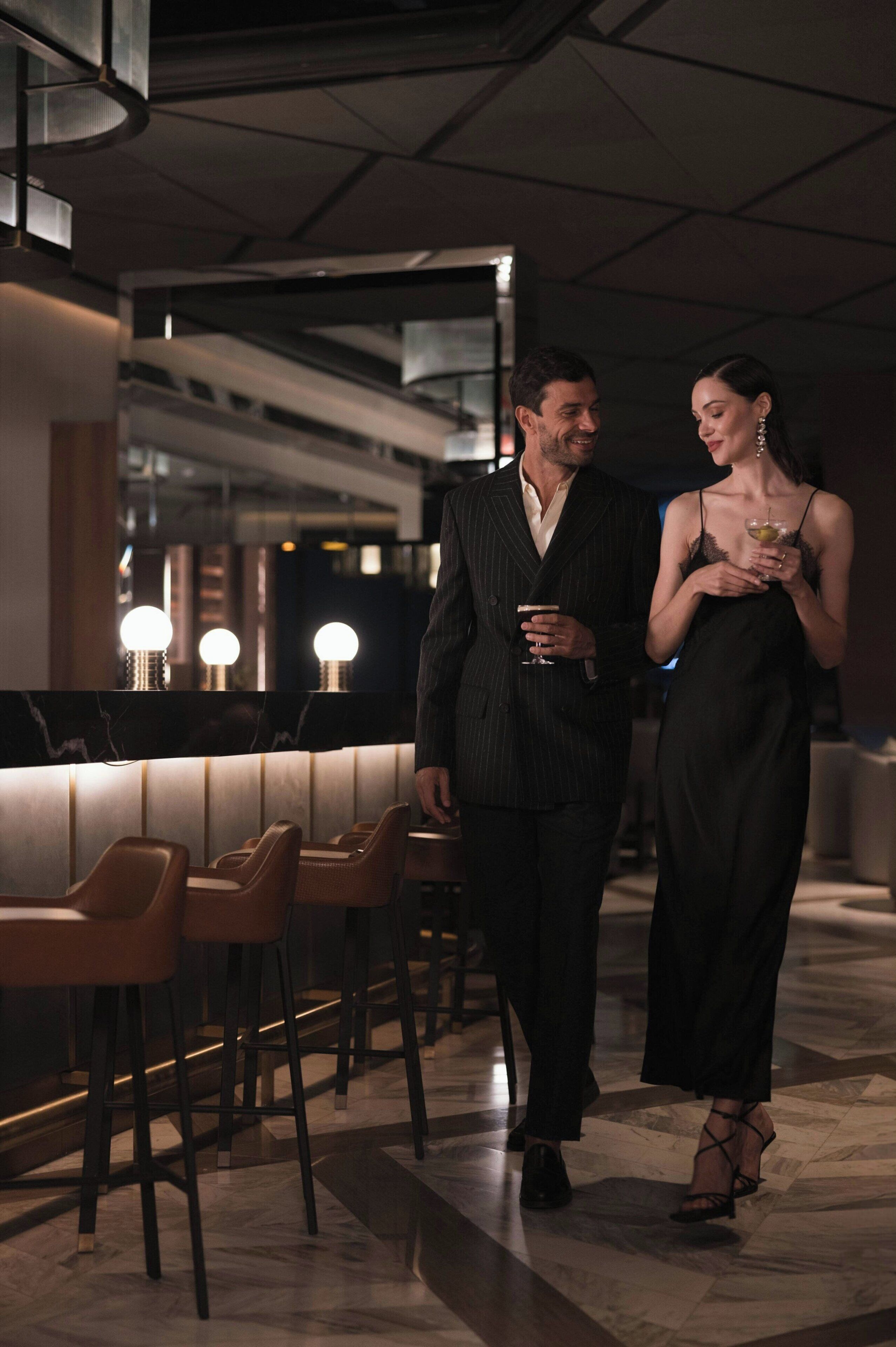 A man and woman walking in a modern bar area with high stools and a marble floor.