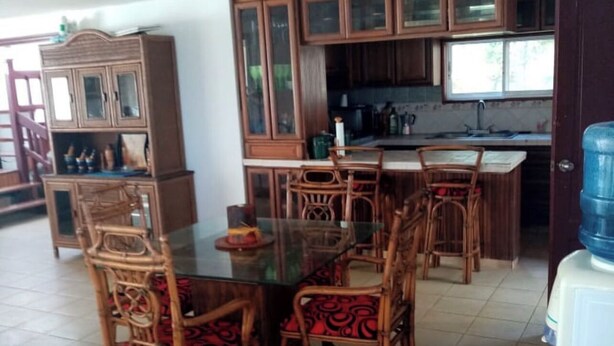 A dining area with a glass-top table surrounded by wicker chairs, wooden cabinets with glass doors, and a kitchen area with a sink and countertop. - Beach house (Cumayasa)
