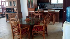 A dining area with a glass-top table and wicker chairs, wooden cabinets, and a water dispenser. - Beach House ( Hidden Paradise  (Cumayasa)