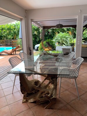 A glass-top dining table with metal chairs, surrounded by a patio area with a view of a swimming pool and lush greenery. - Creole charm with modern comforts (Sainte-Anne)