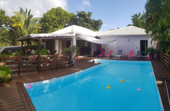 A pool area with a wooden deck, a covered seating area, and a white building with a door. - Creole charm with modern comforts (Sainte-Anne)