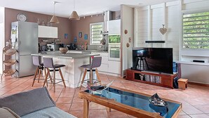 A modern kitchen with a central island, bar stools, a dining table with a glass top, a TV on a wooden stand, and a large window with blinds. - Creole charm with modern comforts (Sainte-Anne)