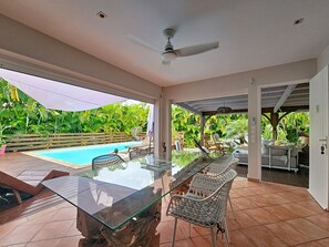 An outdoor patio with a glass-top table and chairs, a ceiling fan, and a view of a swimming pool surrounded by lush greenery. - Creole charm with modern comforts (Sainte-Anne)