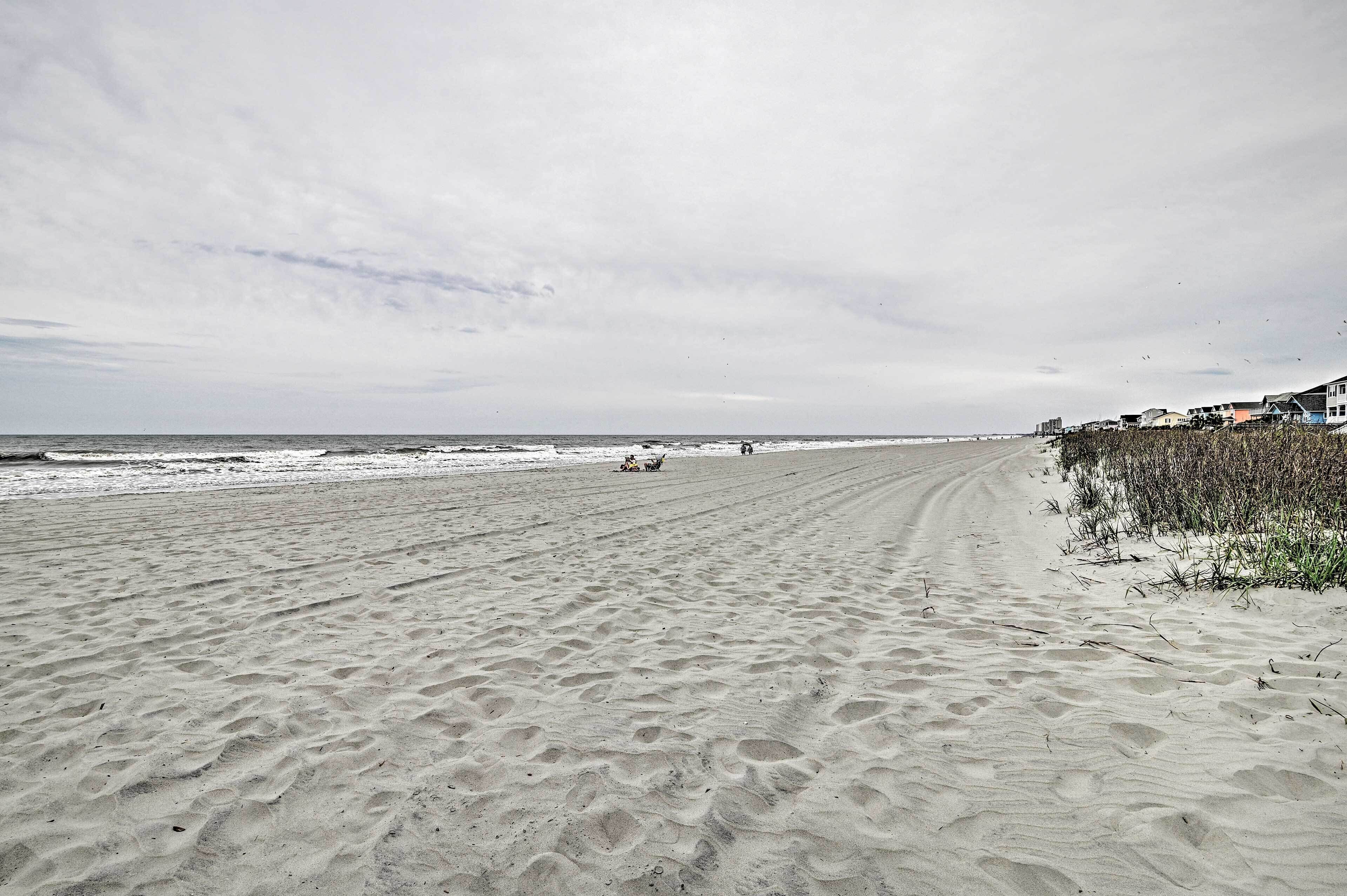 A sandy beach with a clear sky and a few clouds. There are buildings and vegetation near the water's edge.