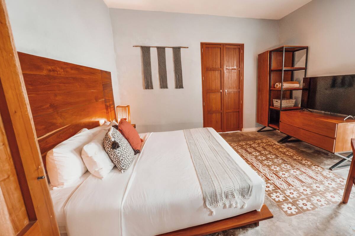 A bedroom with a wooden headboard, a bed with white bedding, a wooden wardrobe, a television, and a patterned rug.