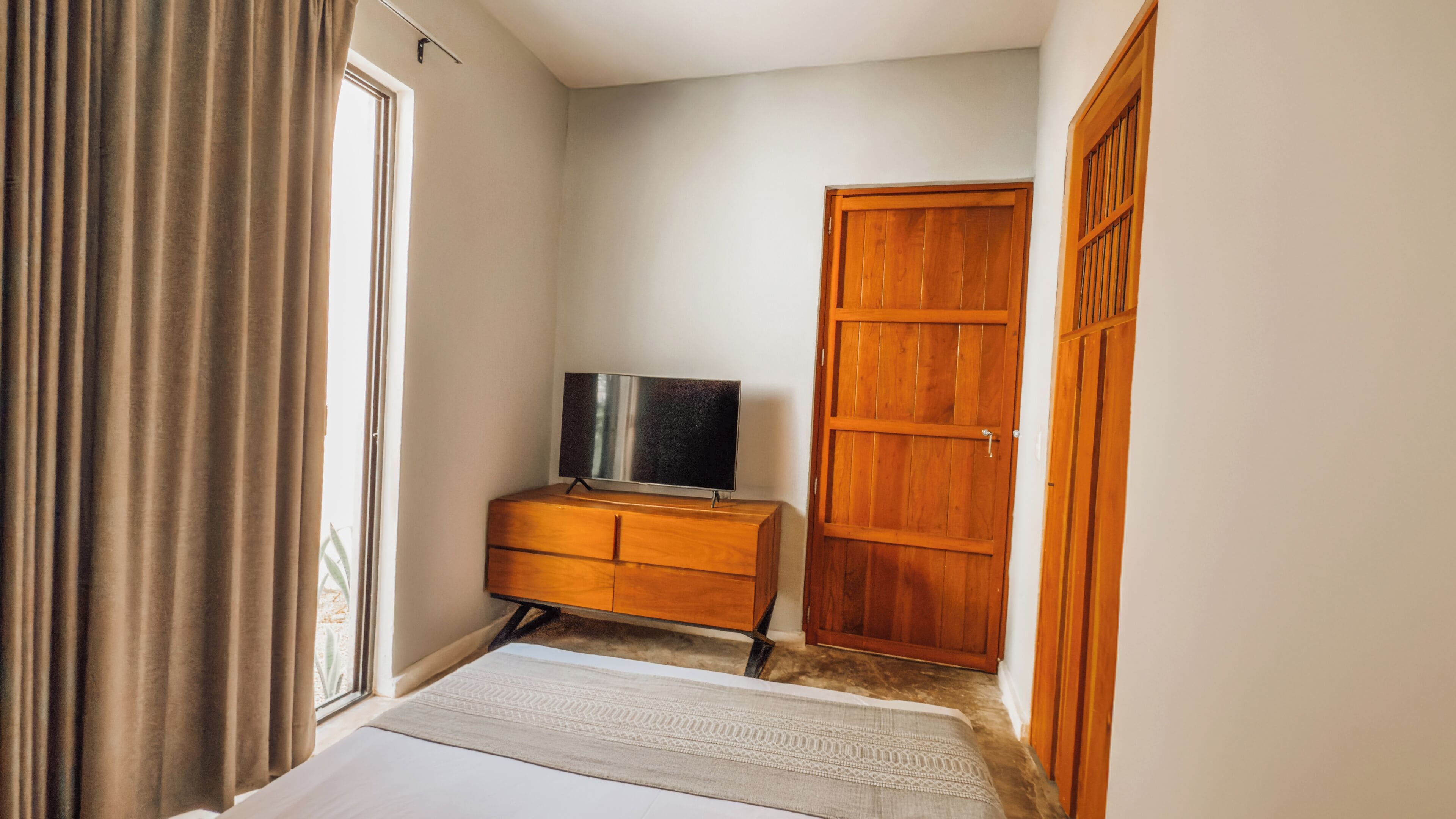 Habitació amb un armari de fusta, una televisió a sobre, una gran porta de fusta i una finestra amb cortines.
