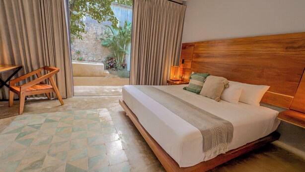 Une chambre d’hôtel avec un grand lit, une tête de lit en bois, une table de chevet avec une lampe, une chaise et une vue sur un jardin par la fenêtre.