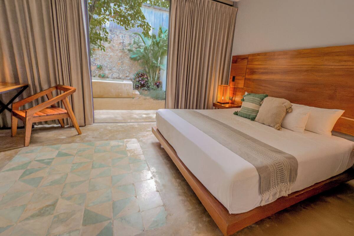 A hotel room with a large bed, a wooden headboard, a nightstand with a lamp, a chair, and a view of a garden through the window.