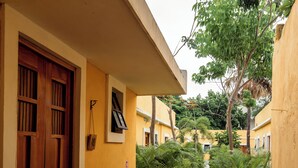 A narrow pathway flanked by lush greenery and yellow buildings with wooden doors.