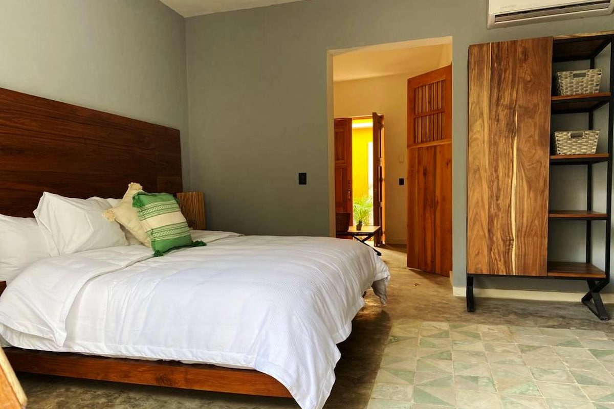 A bedroom with a wooden headboard, a bed with white bedding, a green and white pillow, and a wooden wardrobe with shelves.