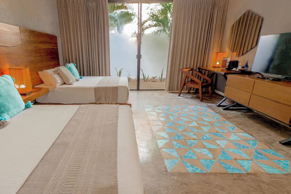 A hotel room with a wooden headboard, two beds with beige and turquoise bedding, a patterned rug, a desk with a chair, and a television.