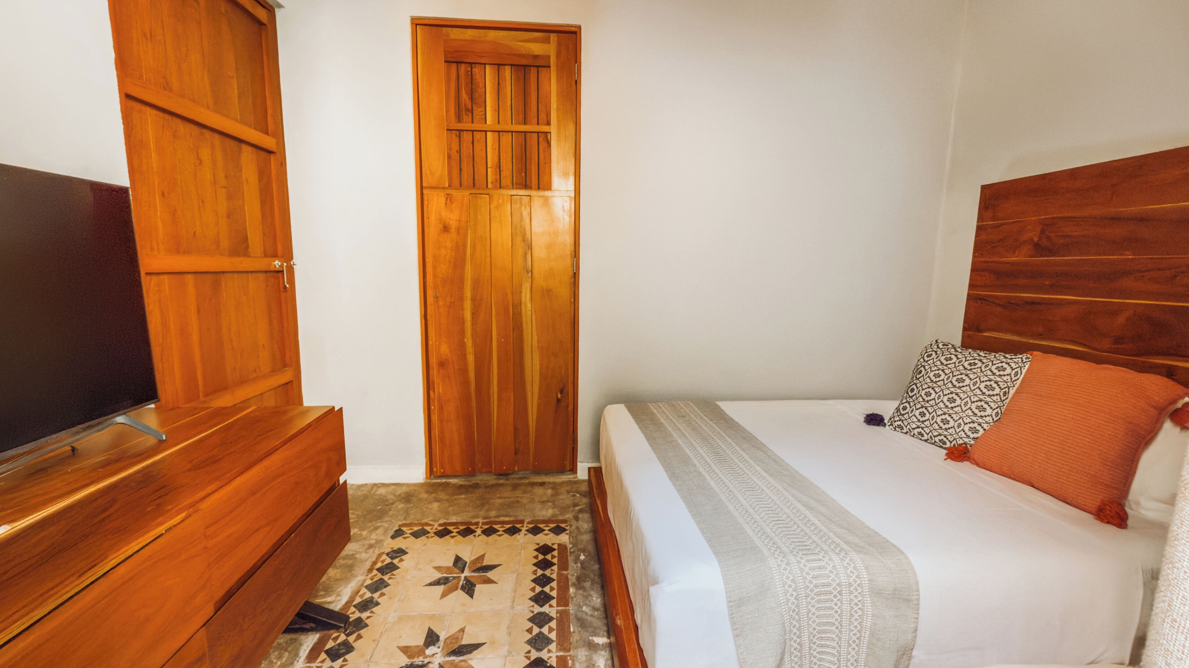 A room with a wooden wardrobe, a television, a bed with patterned pillows, and a tiled floor.
