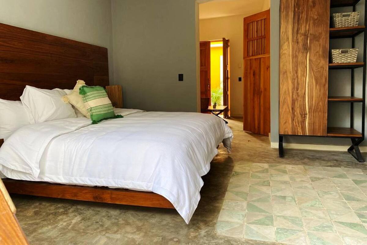 A bedroom with a wooden headboard, a bed with white bedding, a green and white pillow, and a wooden wardrobe with shelves.