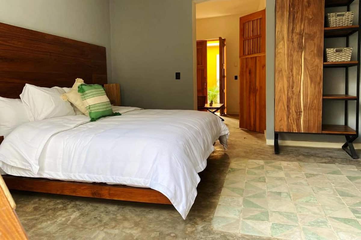Un dormitorio con cabecera de madera, cama con ropa de cama blanca, almohada verde y blanca, y un armario de madera con estantes.