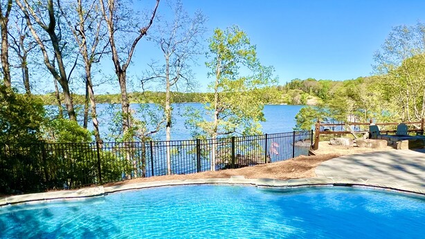 A swimming pool with a stone edge, surrounded by a fence and trees, with a view of a lake and hills in the background. - Massive Lakehouse - Infinity Pool, Hot Tubs, Dock, Firepit (Gainesville)