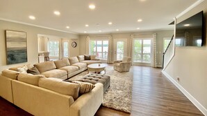 A spacious living room with a large sectional sofa, a round coffee table, and a view of the outdoors through large windows. - Massive Lakehouse - Infinity Pool, Hot Tubs, Dock, Firepit (Gainesville)