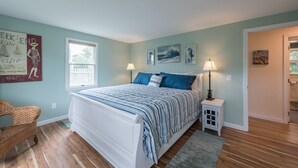Une chambre à coucher avec un lit, deux lampes de chevet, un fauteuil en osier et des œuvres d’art accrochées aux murs.