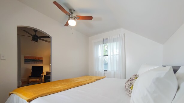 A bedroom with a bed, a ceiling fan, a window with curtains, and a painting on the wall. - Downtown home blocks to the beach w/ king bed, deck fire pit & air conditioning (Manistee)