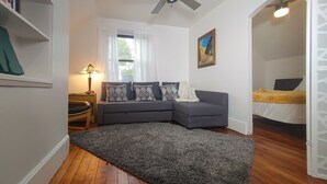 A modern living room with a grey sectional sofa, a patterned rug, a wooden floor, a ceiling fan, a window with white curtains, and a painting on the wall. - Downtown home blocks to the beach w/ king bed, deck fire pit & air conditioning (Manistee)