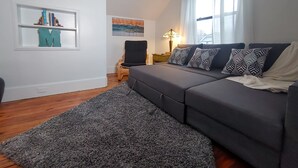 A modern living room with a grey sofa, patterned pillows, a grey rug, a wooden floor, a bookshelf with books and decorative items, a painting on the wall, and a lamp on a side table. - Downtown home blocks to the beach w/ king bed, deck fire pit & air conditioning (Manistee)