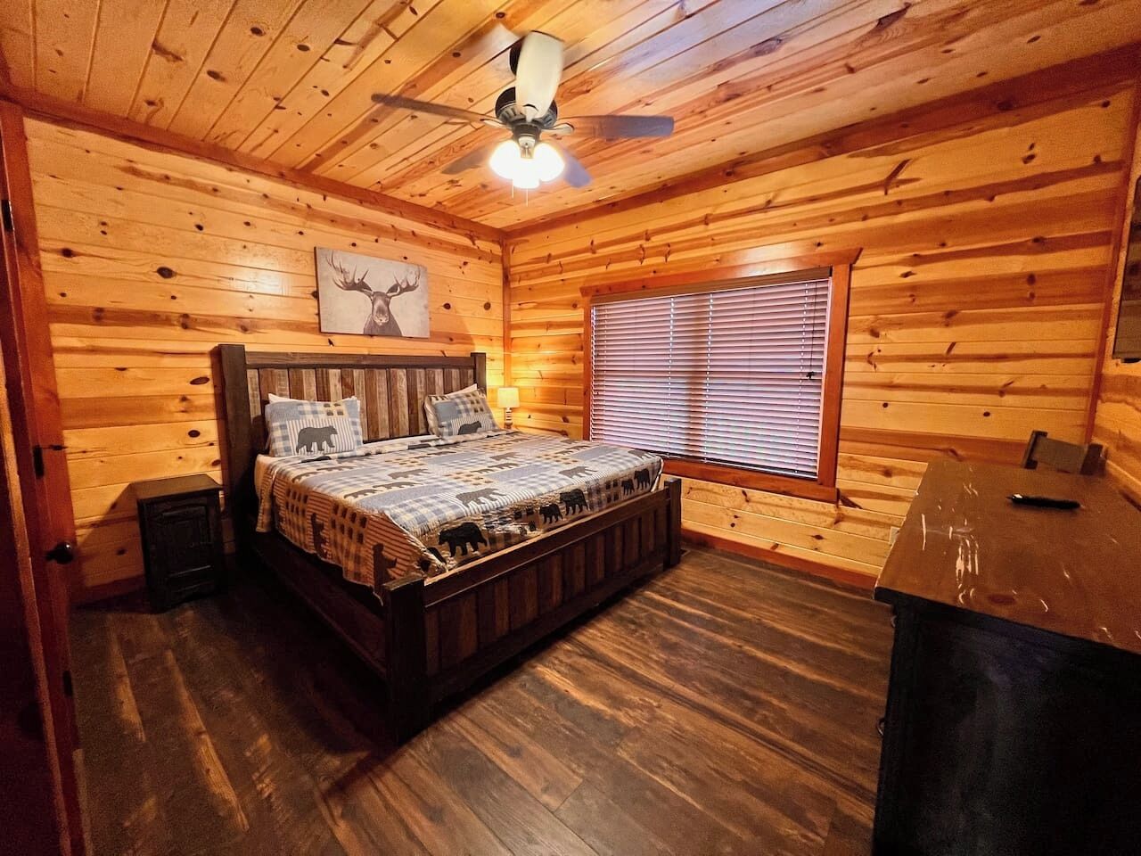 Un dormitorio de cabaña de madera con una cama, un ventilador de techo, una ventana con persianas y una mesita.