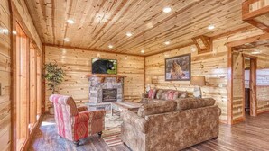 Interior de una cabaña de troncos con chimenea de piedra, sofás de cuero y un televisor montado en la pared.