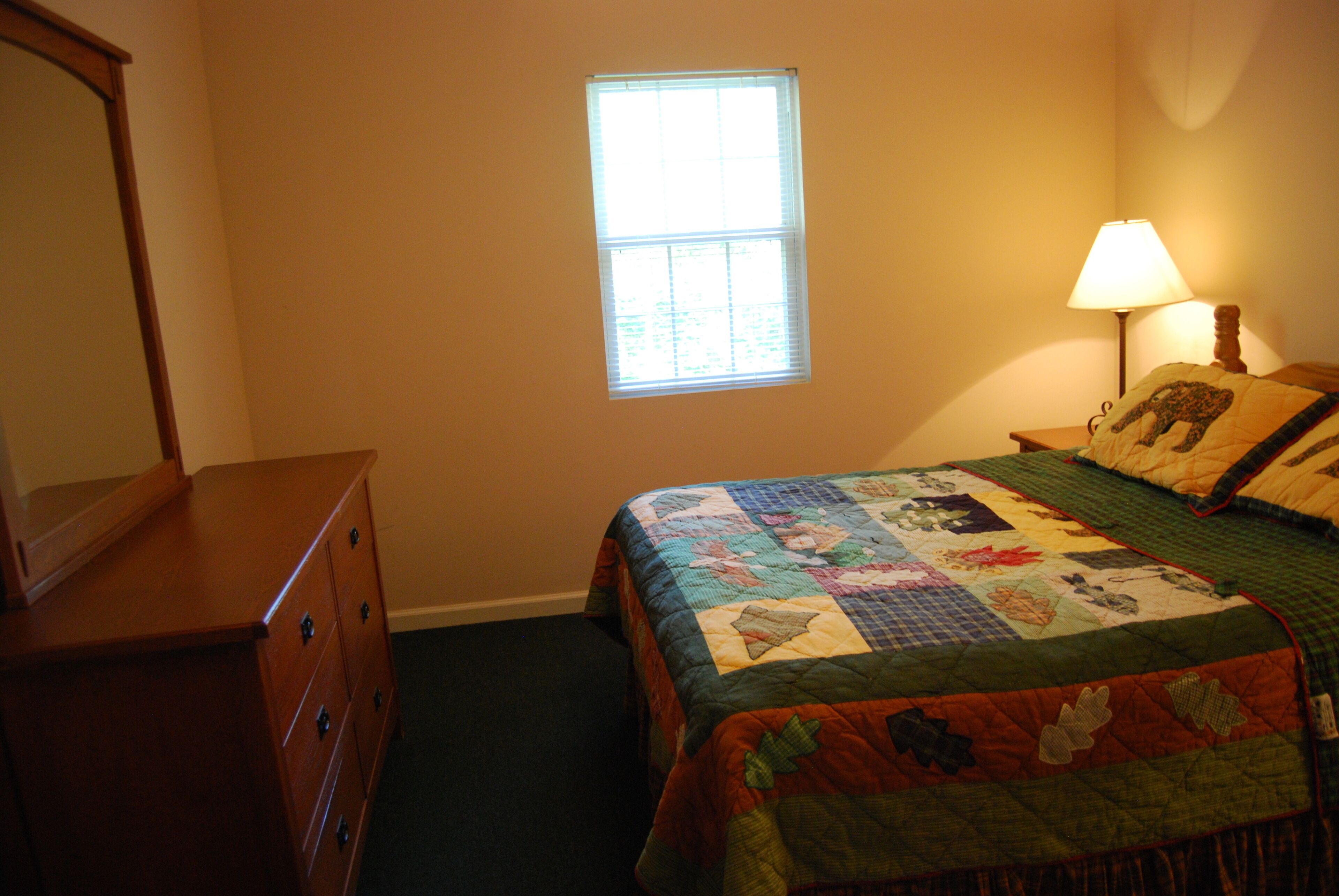A bedroom with a wooden dresser, a quilted bedspread, a window, and a bedside lamp.