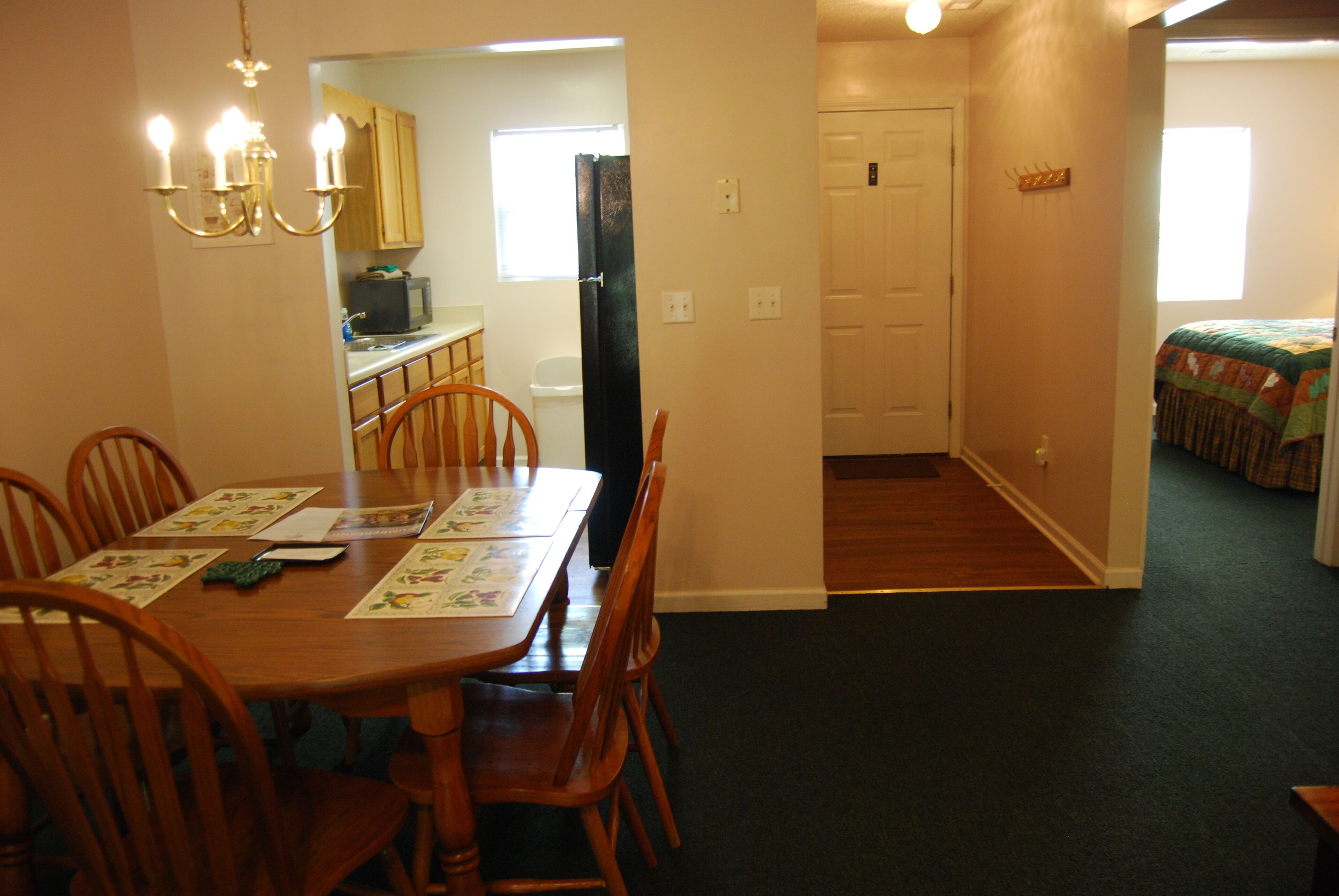 A dining area with a wooden table and chairs, a chandelier, and a kitchen in the background.
