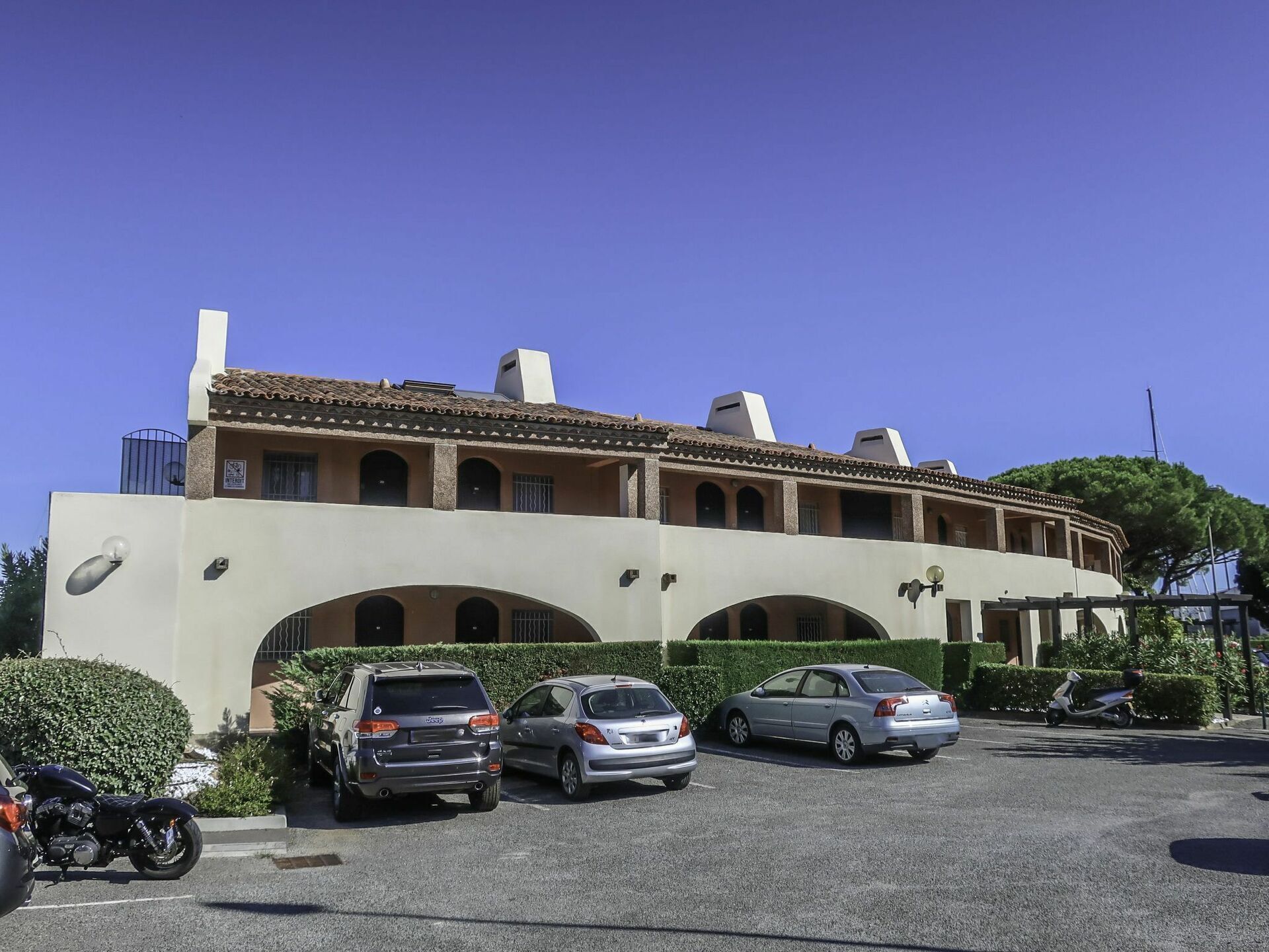 A building with a beige facade, multiple arches, and a tiled roof. There are three parked cars and a motorcycle in front.
