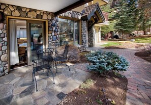 A stone exterior with a patio area featuring two metal chairs and a small evergreen tree.