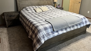 Een slaapkamer met een houten hoofdbord, een geruit beddegoed, een nachtkastje met een lamp en een stapel boeken.
